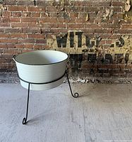 Large cream-colored metal bin with concentric ridges on sides sitting on a tripod black metal stand with curled leg ends, placed indoors against an aged brick wall.