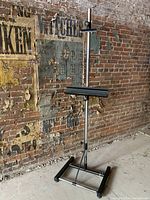 Full view of large professional modern artist's easel against a brick wall background, showing metal construction and adjustable features.