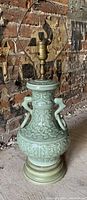 Tall celadon porcelain vintage table lamp with double handles, intricate raised design and wooden base, photographed against a rustic brick wall background.