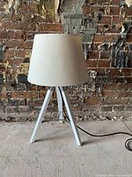 Photo of modern tripod lamp with painted wood legs and white fabric shade against a distressed brick wall background.
