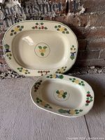 Front view of two ceramic platters with fruit painting on cream ceramic, displayed against brick wall.