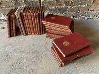 Full group photo of 25 red leather bound books with gilt decorated spines and covers arranged in two stacks on wooden surface against brick wall.