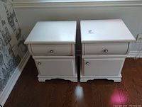 Two white wooden night tables side by side showing front and side views with drawer and cabinet door, scuffed and worn, placed on hardwood floor against wall with wallpaper.