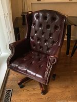 Front view of burgundy faux leather executive office chair showing button tufted backrest and seat, nailhead trim, and wood-tone base with caster wheels.