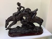 Side view of bronze-tone horse statue with two jockey riders jumping over a hurdle on a wooden base, showing detailed features and overall condition.