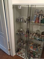 Glass IKEA display cabinet shelf unit with clear glass panels and white frame. Shelves and door visible with reflections. Cabinet filled with snow globes and other items (not included).