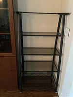 Front angled view of the bookshelf showing four smoked tempered glass shelves on a black metal frame with wood accents on top corners.