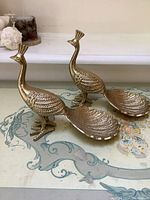 Pair of gold-painted metal peacock figurines with tail feathers splayed out to form small trays, on a decorative surface, showing front and left side.