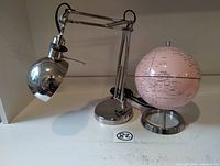 Silver articulated desk lamp next to a pink desk globe both on a white surface.