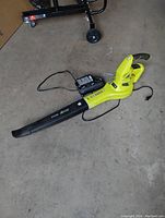 RYOBI 18V hybrid leaf blower lying on concrete floor with battery charger and power cord. Good condition showing minor cosmetic wear.