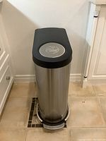 Front angled view of the stainless steel trash can with black lid and foot pedal on beige tile floor.