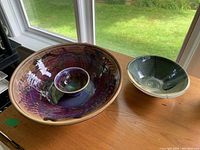 Photo showing a large purple and reddish glazed pottery bowl and a medium blue and beige glazed pottery bowl side by side on a wooden surface with grass background.