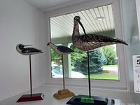 Three hand painted bird sculptures on stands shown by window, heights from 13 to 17 inches, one missing beak.