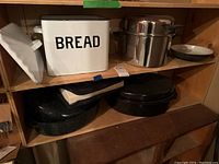 Shelf with white enamel bread box labeled 'BREAD', large stainless steel stock pot with lid, two black enamel roasting pans, and vintage enamel bowl.