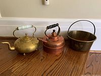 Photo shows three antique metal items on wooden floor near baseboard: brass teapot with white handle, Simplex copper teapot with black handle, and a brass pot with a large handle, highlighting their shape, size and condition.