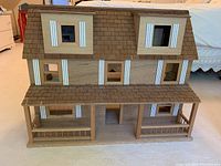 Front exterior view of the wooden doll house showing the brown shingled roof, multiple windows with white trim, and front porch with railing.