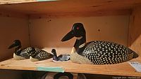 Three handcrafted hand-painted loon figurines arranged on a wooden shelf, varying in size.