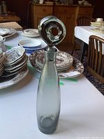 Full view of the Holmegaard glass decanter with its circular hole stopper on a table with plates and dishes in the background.