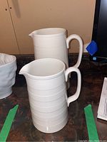 Two white ironstone pitchers placed side by side on a dark surface, showing size difference and ribbed pattern detail.