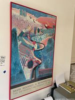 Framed colorful landscape art print with red frame leaning on a beige wall.