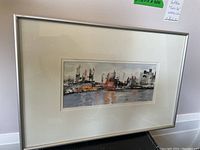 Framed landscape watercolour painting showing a city skyline with water reflections, framed in a silver-colored frame with white matting.