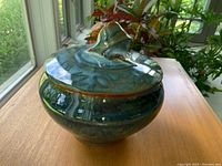 Closed pottery bowl with blue-green glaze and decorative lid handle, set on wooden table near window.