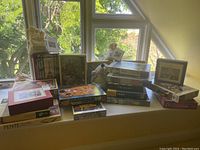 Wide photo showing multiple puzzle boxes stacked on window ledge including hand cut wooden puzzles and Eurographics puzzles, plus a stuffed animal dog plush.