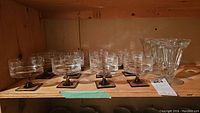 Twelve Rosenthal glasses with clear bowls and smoky glass stems with square bases arranged on a wooden shelf.