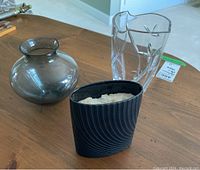 Three vases on wooden table including dark grey round vase, clear etched glass vase, and black textured Rosenthal Drift Lava vase stuffed with packing foam.
