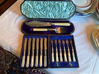 Top-down image showing two cutlery sets in open cases, fish serving set on top with satin blue lining and dessert set below with velvet blue lining