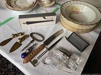 Photo showing six Sovereign Potters plates stacked, decorative brass knives, corkscrew, blue art glass stopper, boxed wine finer, letter opener, and clear acrylic holder.