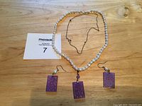 Full view of pearl necklace with sterling chain and matching earrings with purple carved rectangular pendants arranged on wooden surface.