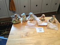 Five Calico cat statues arranged on wooden table near kitchen cabinets. Three statues partially wrapped in clear plastic. Cat statues are in various clothing and poses.