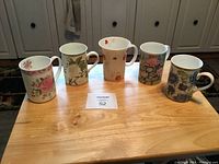 Five vintage china coffee mugs with different floral designs arranged in a row on a wooden table, showing their side views and handles.