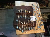 Full view of wooden display rack with mounted collector's spoons and loose spoons and fork beside it on table