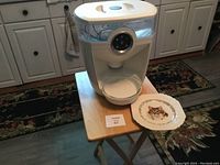 White automatic food dispenser with WiFi and sensor, placed on a small wooden foldable table next to the cat-themed ceramic plate.
