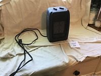 Full view of mini black electric heater with attached cord, placed on cloth surface.