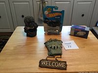 Full view of table with all items: Hot Wheels Batman car in original packaging, black gorilla ashtray, green mailbox decor, wooden Welcome sign on chain