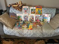 Wide shot showing entire collection of children's books on couch including various yearbooks and colorful character storybooks.