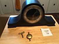 Front view of black wooden mantle clock with curved top and round clock face on wooden table with winding key and pendulum in front.