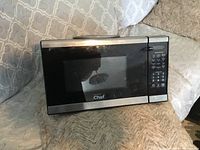 Front and side view of the stainless steel Master Chef microwave oven placed on a beige couch showing the door, digital display and control pad.