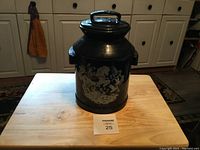 Front view of large black ceramic cookie jar shaped like milk can with white floral design on front and handle on lid, sitting on wooden table