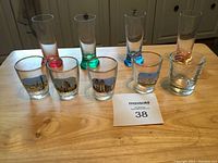 Photo of eight assorted shot and B52 glasses on a wooden table with a MaxSold tag numbered 38.