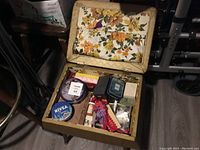 Top view of open sewing stool with floral upholstery inside containing tin containers, sewing threads, lace trims, and other assorted sewing accessories.