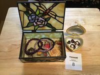 Open rectangular stained glass jewelry box containing bracelets and charms on fabric lining and a small open heart-shaped box with rings and earrings beside it on a wooden table.