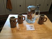 Four beer steins on a wooden table, three ceramic with raised reliefs and one glass with pewter lid