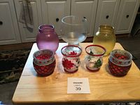 Seven small decorative items arranged on a wooden table including frosted purple and yellow glass vases, a large clear vase, two red glass holders with silver and bead embellishments, and two small clear glass holders painted with holiday designs.