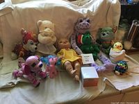 Collection of assorted vintage toys arranged on a beige cloth, showing plush animals, dolls, and figurines in used condition.