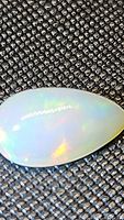 Close-up image of pear-shaped Ethiopian opal cabochon gemstone showing rainbow flashes over milky white base color on textured black fabric.