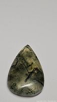 Clear image of pear-shaped mossy green prehnite cabochon with black rutilated needle inclusions on plain background.